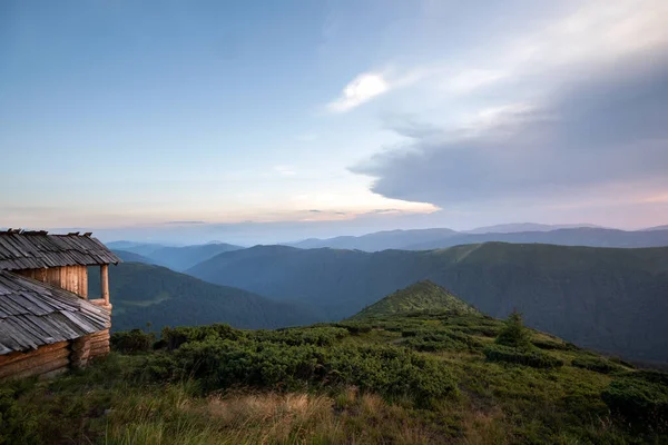 Verão paisagem montanhosa noite com velho abrigo turístico abandonado em colinas gramíneas e picos distantes ao pôr do sol colorido — Fotografia de Stock