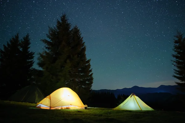 Tentes de camping lumineuses qui brillent sur le camping dans les montagnes sombres sous le ciel étoilé couvert de nuit. Style de vie actif et concept de voyage — Photo