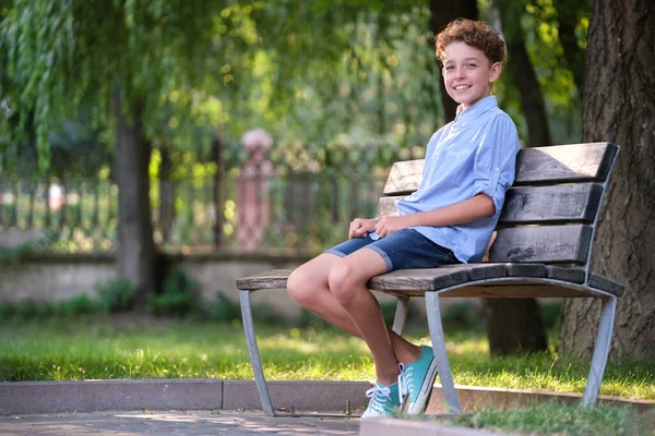 Young happy child boy relaxing sitting on bench in summer park. Positive kid enjoying summertime outdoors. Child wellbeing concept — Stock Photo, Image