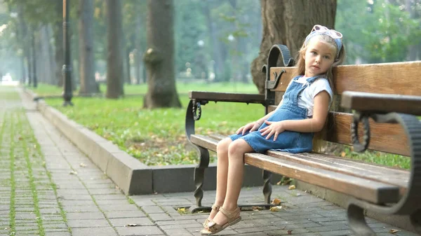 Small happy child girl sitting on a bench resting in summer park — Stock Photo, Image