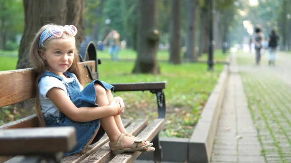 Pequeña niña feliz sentada en un banco descansando en el parque de verano —  Fotos de Stock