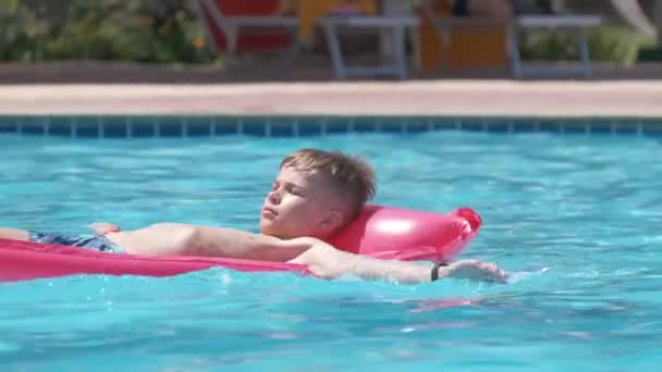 Молодой радостный мальчик весело плавает на надувном надувном матрасе в бассейне с голубой водой в теплый летний день на тропических каникулах. Концепция летних мероприятий — стоковое видео