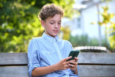 Yaz parkında akıllı telefonuyla oyun oynayan genç bir çocuk. Elektronik cihazlar konseptinden bağımlılık