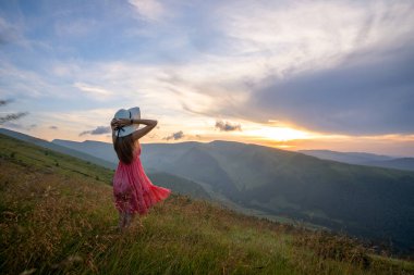 Kırmızılı genç kadın yaz dağlarında rüzgarlı bir akşamda yamaçtaki çayırda yürüyor.