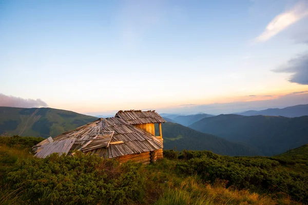 Verão paisagem montanhosa noite com velho abrigo turístico abandonado em colinas gramíneas e picos distantes ao pôr do sol colorido — Fotografia de Stock