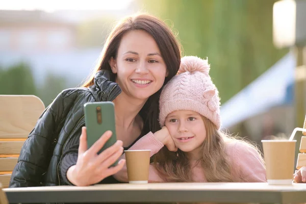 젊은 엄마와 딸은 화창 한 가을 날 거리 카페에서 뜨거운 음료를 마시며 휴대폰 셀카 카메라로 사진을 찍고 있다. 일상 생활 에서의 소셜 미디어의 존재 — 스톡 사진