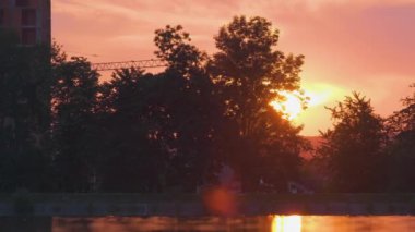 Göl suyuna yansıyan karanlık park ağaçlarının gölgeli manzarası ve gün batımında parkın tepesinde yürüyen yayalar.