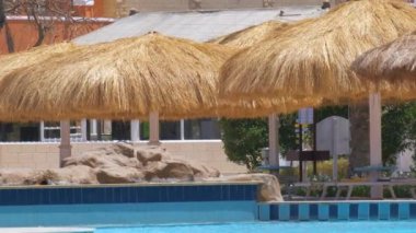 Boş güverte sandalyeleri, tropik tatil köylerinde havuz kenarında saman gölgesi şemsiyelerinin altında. Yaz tatilleri ve kaçış konsepti