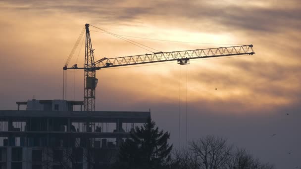 Dark silhouette of tower crane at high residential apartment buildings construction site at sunset. Real estate development. — Stock Video