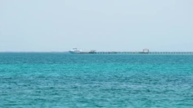 Mavi deniz suyunun dalgalı yüzeyi ve sakin dalgalar üzerinde yüzen uzun rıhtımın sonundaki uzak gemiyle deniz manzarası.