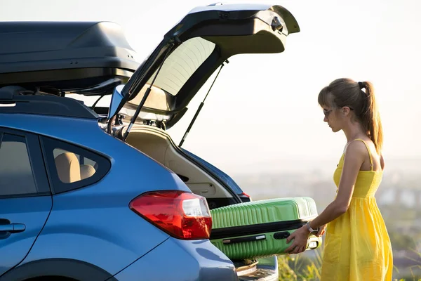 Conductora en vestido de verano poniendo maleta verde dentro del maletero de su coche. Concepto de viajes y vacaciones — Foto de Stock