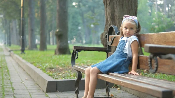 Little child girl sitting alone on a bench in summer park — Stock Photo, Image