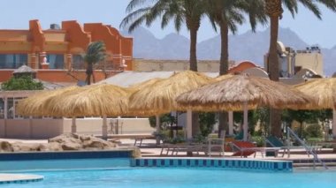 Boş güverte sandalyeleri, tropik tatil köylerinde havuz kenarında saman gölgesi şemsiyelerinin altında. Yaz tatilleri ve kaçış konsepti