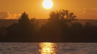 Göl suyuna yansıyan karanlık park ağaçlarının gölgeli manzarası ve gün batımında parkın tepesinde yürüyen yayalar.