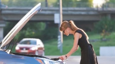 Kafası karışmış bir kadın sürücü arabasının yanındaki caddede durmuş kaputu patlamış ve bozuk motora bakıyor.