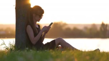 Genç bir kadın, göl kıyısındaki çimlerin üzerinde oturmuş sıcak bir yaz akşamında cep telefonunu karıştırıyor. İletişim ve mobil bağlantı kavramı