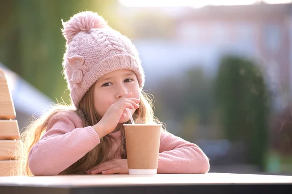 stock image Portrait of cute little child girl in pink hat sitting alone at street cafe drinking tea from paper cup