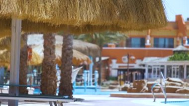Boş güverte sandalyeleri, tropik tatil köylerinde havuz kenarında saman gölgesi şemsiyelerinin altında. Yaz tatilleri ve kaçış konsepti