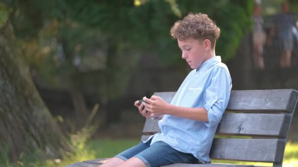 Νεαρό αγόρι παίζει παιχνίδι στο smartphone του σε εξωτερικούς χώρους στο καλοκαιρινό πάρκο. Εθισμός από την έννοια των ηλεκτρονικών gadgets — Αρχείο Βίντεο