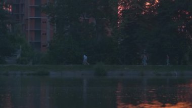 Göl suyuna yansıyan karanlık park ağaçlarının gölgeli manzarası ve gün batımında parkın tepesinde yürüyen yayalar.