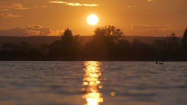 Göl suyunda kırılan park ağaçlarından oluşan karanlık bir siluet ve gün batımında parkın tepesinde yürüyen yayalar ile Lakeside manzarası