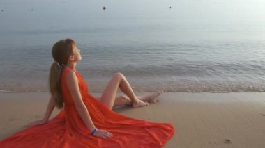 Deniz kıyısında dinlenen kırmızı elbiseli mutlu genç kadın sıcak yaz sabahının tadını çıkarıyor.
