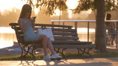 Parktaki bankta oturan genç bir kadın sıcak bir yaz akşamında cep telefonunu karıştırıyor. İletişim ve mobil bağlantı kavramı