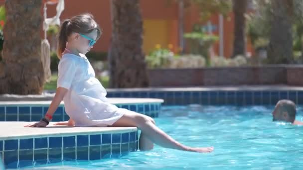 En ung, glad jente som hviler på bassengsiden med klart blått vann på solrik sommerdag. Begrepet tropiske ferier – stockvideo