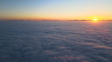 Beyaz, yoğun, sisli bulutların üzerindeki renkli günbatımının havadan görünüşü ufuktaki dağ tepelerinin uzak karanlık siluetleriyle kaplıdır.