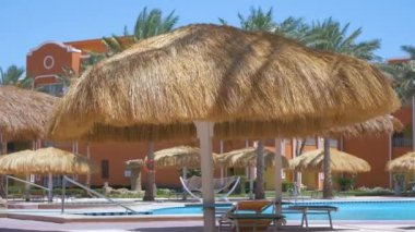 Boş güverte sandalyeleri, tropik tatil köylerinde havuz kenarında saman gölgesi şemsiyelerinin altında. Yaz tatilleri ve kaçış konsepti