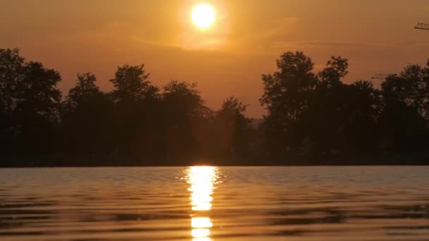 湖の水に映し出された公園の木々の暗いシルエットと湖畔の風景と明るい夕日で堤防上の遠く歩行者の人々 — ストック動画