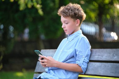 Yaz parkında akıllı telefonuyla oyun oynayan genç bir çocuk. Elektronik cihazlar konseptinden bağımlılık