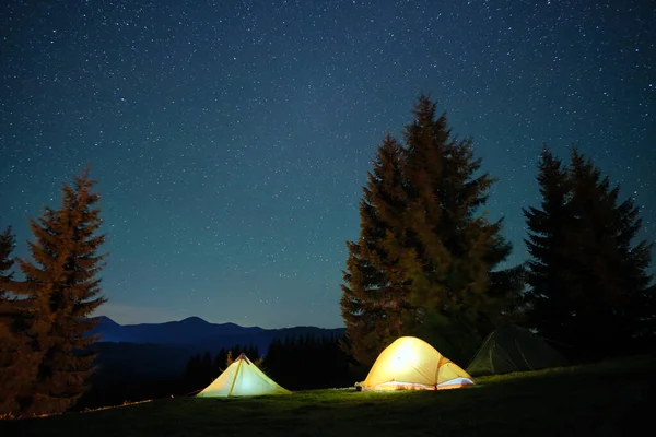 Des tentes touristiques lumineuses illuminées brillent sur le camping dans les montagnes sombres sous le ciel nocturne avec des étoiles scintillantes. Concept de mode de vie actif — Photo