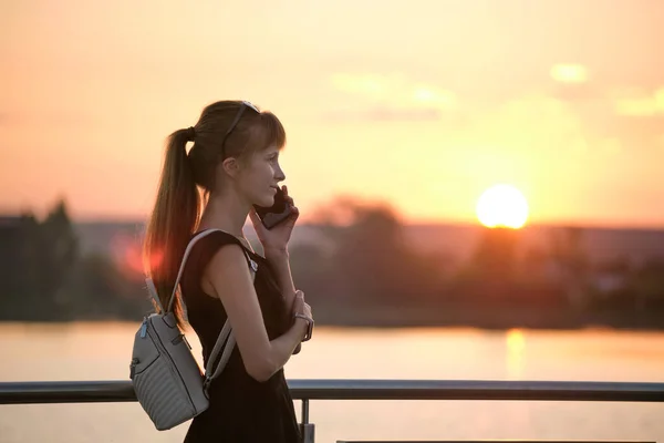 Счастливая молодая женщина отдыхает на берегу озера в летнем парке, разговаривая по мобильному телефону на открытом воздухе в теплый вечер. Концепция связи и мобильной связи — стоковое фото