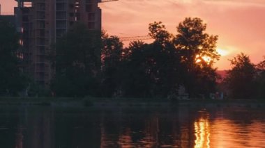 Göl suyuna yansıyan karanlık bina silüetiyle Lakeside manzarası ve parlak günbatımında toprak üzerinde yürüyen yayalar.