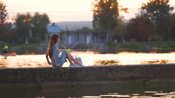 Visão traseira da jovem mulher solitária sentada sozinha na margem do lago desfrutando de noite quente. Bem-estar e relaxamento no conceito de natureza — Vídeo de Stock
