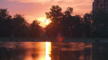 Göl suyunda kırılan park ağaçlarından oluşan karanlık bir siluet ve gün batımında parkın tepesinde yürüyen yayalar ile Lakeside manzarası