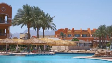 Boş güverte sandalyeleri, tropik tatil köylerinde havuz kenarında saman gölgesi şemsiyelerinin altında. Yaz tatilleri ve kaçış konsepti