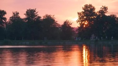 Göl suyunda kırılan park ağaçlarından oluşan karanlık bir siluet ve gün batımında parkın tepesinde yürüyen yayalar ile Lakeside manzarası