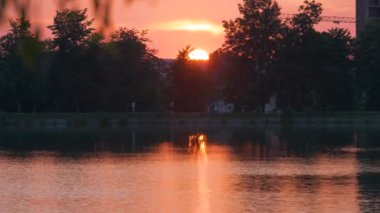 Göl suyunda kırılan park ağaçlarından oluşan karanlık bir siluet ve gün batımında parkın tepesinde yürüyen yayalar ile Lakeside manzarası