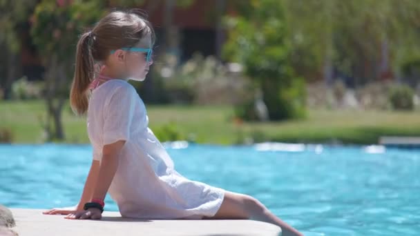 Portrait d'enfant heureuse fille en robe blanche relaxant sur le côté de la piscine le jour ensoleillé d'été pendant les vacances tropicales — Video
