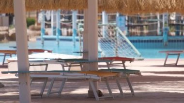 Boş güverte sandalyeleri, tropik tatil köylerinde havuz kenarında saman gölgesi şemsiyelerinin altında. Yaz tatilleri ve kaçış konsepti