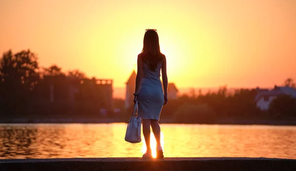 Back view of lonely young woman standing alone on lake shore enjoying warm evening. Wellbeing and relaxing in nature concept — Stock Photo, Image