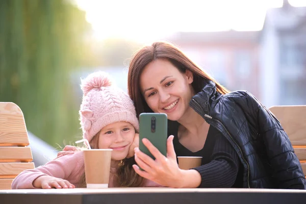 아름다운 한 젊은 여성 이 밝은 가을에 딸 과 함께 카페에서 뜨거운 음료를 마시며 핸드폰 셀카 카메라로 사진을 찍고 있습니다. 가족 관계 에서의 행복 — 스톡 사진