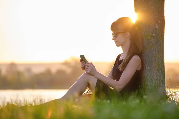 Молодая женщина, сидящая на зеленой газоне на берегу озера, просматривает свой мобильный телефон на открытом воздухе теплым летним вечером. Концепция связи и мобильной связи — стоковое фото