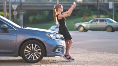 Arabasının yanında duran genç bir kadın sürücü yazın bir şehir caddesinde cep telefonuyla konuşuyor.