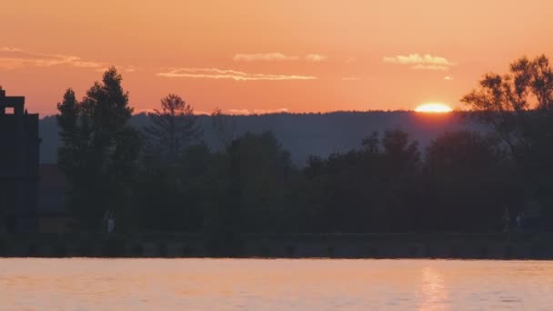 Пейзаж на березі озера з темним силуетом паркових дерев, відображених у воді озера та віддалених пішохідних людей на набережній на яскравому заході сонця — стокове відео