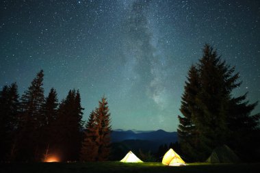 Gece gökyüzünün altındaki karanlık dağ ormanlarında, parlayan yıldızlarla parlayan kamp alanının yakınında parlak turist çadırları. Aktif yaşam tarzı ve açık hava yaşam konsepti