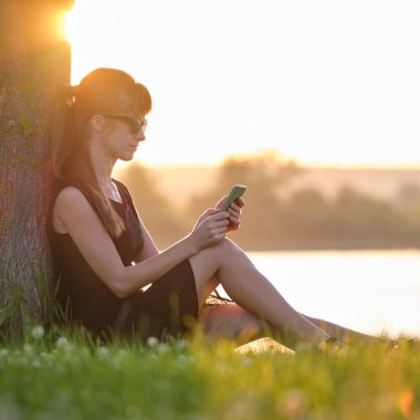 Genç bir kadın göl kenarındaki çimenlerde rahat rahat cep telefonuna bakıyor ılık bir yaz akşamında. İletişim ve mobil bağlantı kavramı
