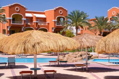 Boş güverte sandalyeleri, tropik tatil köylerinde havuz kenarında saman gölgesi şemsiyelerinin altında. Yaz tatilleri ve kaçış konsepti